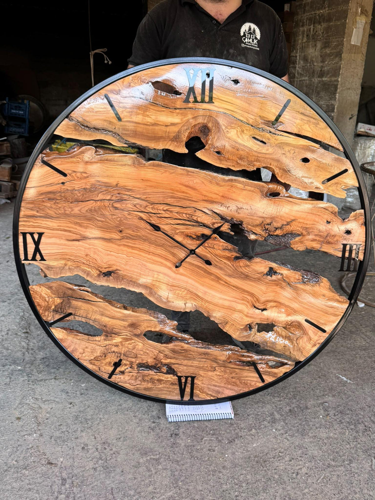 Extra large handmade olive wood wall clock front-on in the workshop, with black metal ring and Roman numerals