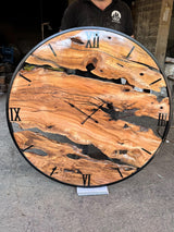 Extra large handmade olive wood wall clock front-on in the workshop, with black metal ring and Roman numerals